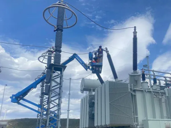Técnico en canastilla trabajando junto a las boquillas de un transformador de potencia, con la estructura del sistema de protección contra descargas en primer plano