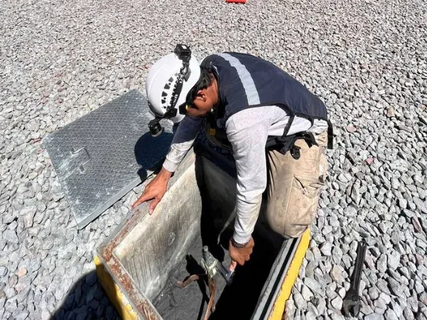 Técnico levanta la tapa del registro de inspección de la malla de tierras en el patio de grava de una subestación