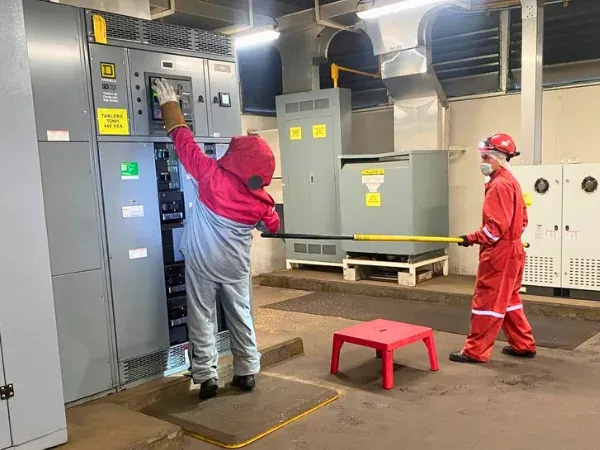Trabajador con traje completo para arco eléctrico maniobrando el interruptor principal de un tablero de 480 V sobre tapete dieléctrico, con un compañero de respaldo a distancia