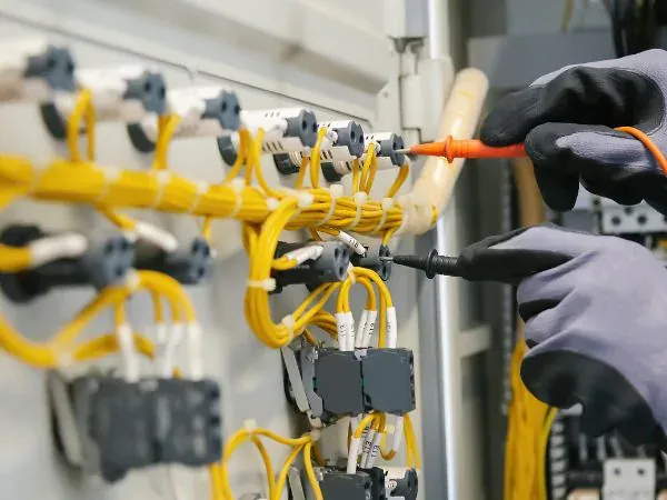 Manos con guantes dieléctricos midiendo con puntas de multímetro sobre el cableado de control amarillo y las borneras de un tablero eléctrico