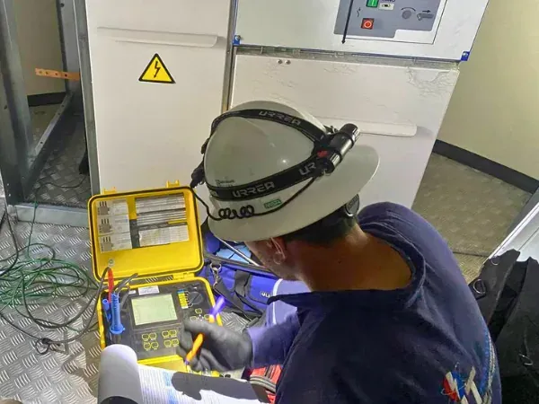 Técnico con casco y lámpara frontal tomando lecturas de un instrumento de prueba portátil frente a una celda de media tensión