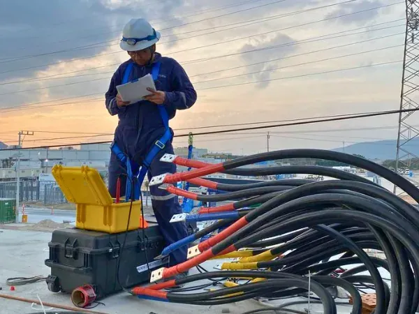 Ingeniero registrando lecturas junto a un equipo de prueba con múltiples conductores conectados, en el patio de una instalación eléctrica industrial