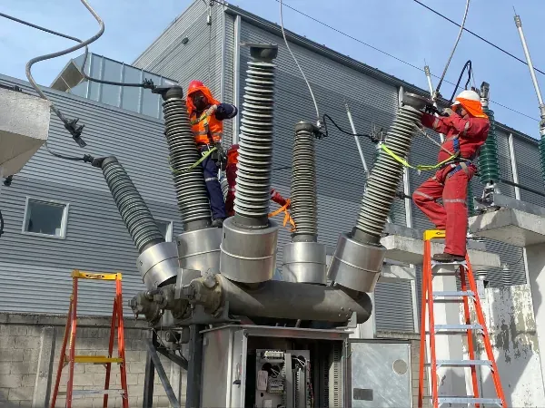 Dos técnicos con casco y arnés trabajan sobre las columnas de aisladores de un seccionador de alta tensión en la subestación de una planta
