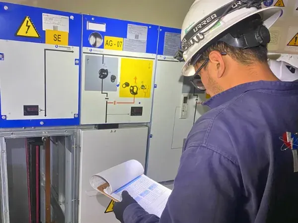Ingeniero con el protocolo de pruebas en mano frente al frente de maniobra de un tablero de media tensión con diagrama mímico y señalización de riesgo eléctrico
