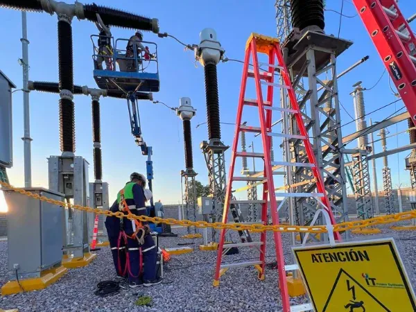 Técnicos en canastilla elevadora interviniendo las boquillas de un equipo de alta tensión en una subestación de intemperie delimitada con señalización de seguridad